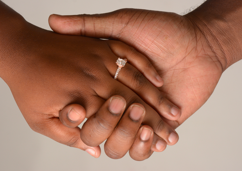 A black male hand holding a black female hand with the female hand turned out to showcase an engagement ring.
