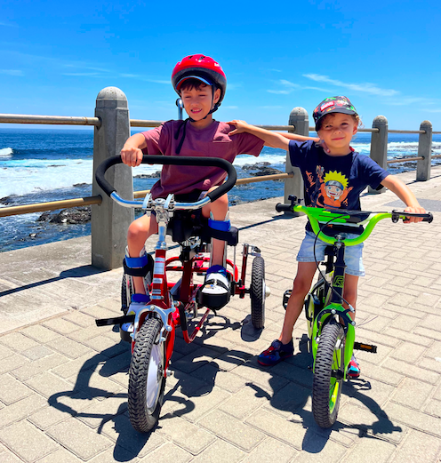 Elijah on a red adapted bike with his brother, Isaiah, on a green bicycle on a promenade next to the ocean
