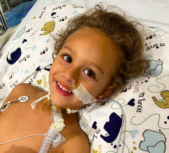 Elijah Cottle in a hospital bed with tubes in his nose and throat to assist with breathing. He is smiling in the photo