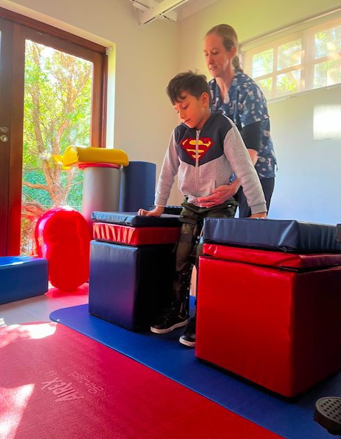 Elijah during therapy, standing between two padded pillars with his therapist assisting him to stand.