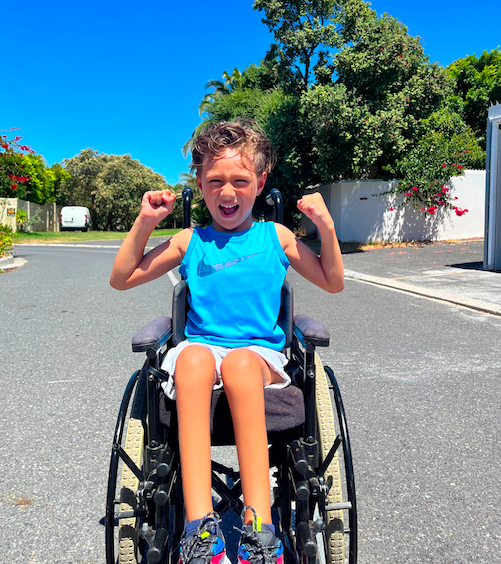 Elijah outside on the road in the sunshine wearing a bright blue shirt in his wheelchair, showing off his muscles