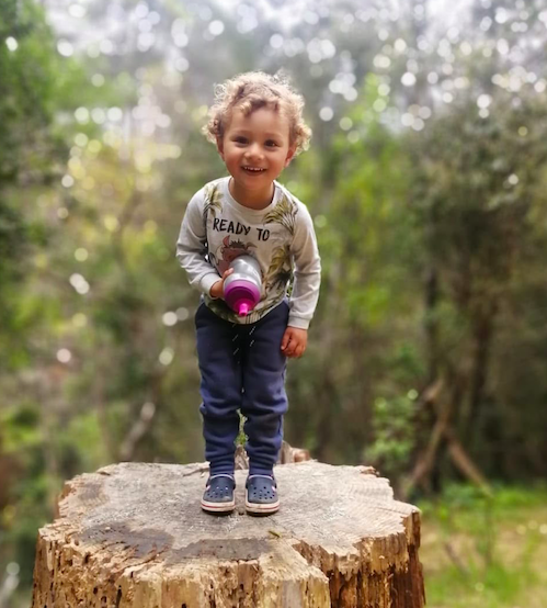 Elijah Cottle as a toddler standing on a tree stump with his brown hair, tanned skin, grey shirt, jeans and trainers